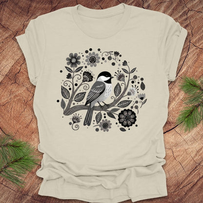 Beige t-shirt of a chickadee on a branch surrounded by folk art flowers, all in black and white, on a wooden background.