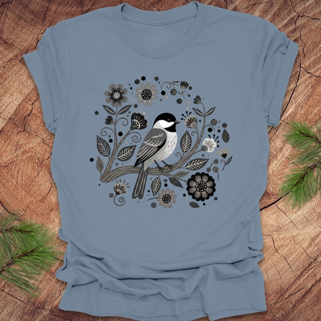 Steel blue t-shirt of a chickadee on a branch surrounded by folk art flowers, all in black and white, on a wooden background.