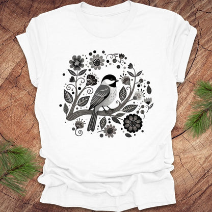 White t-shirt of a chickadee on a branch surrounded by folk art flowers, all in black and white, on a wooden background.