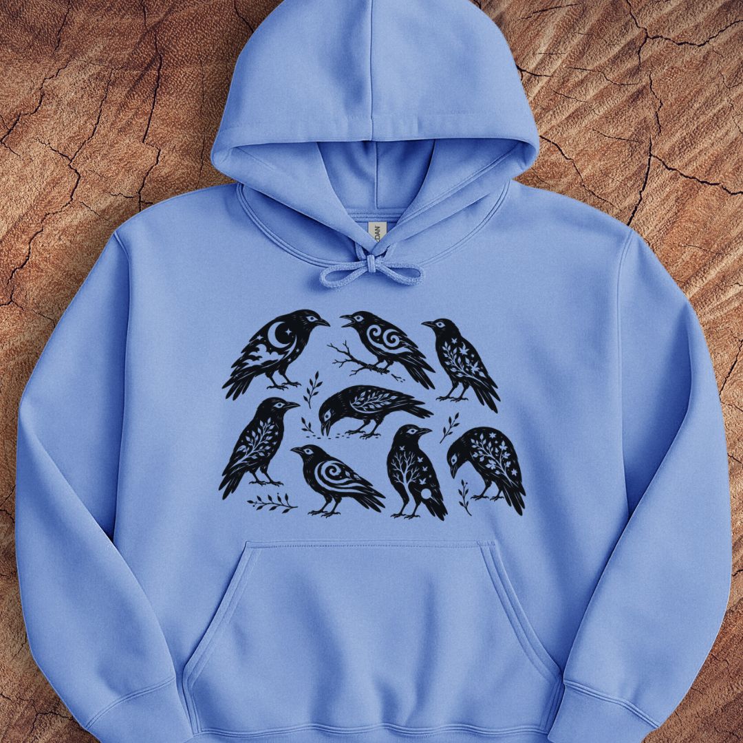 A light blue pullover hoodie with a black graphic print of seven stylized birds, laid flat on a textured brown wood background.