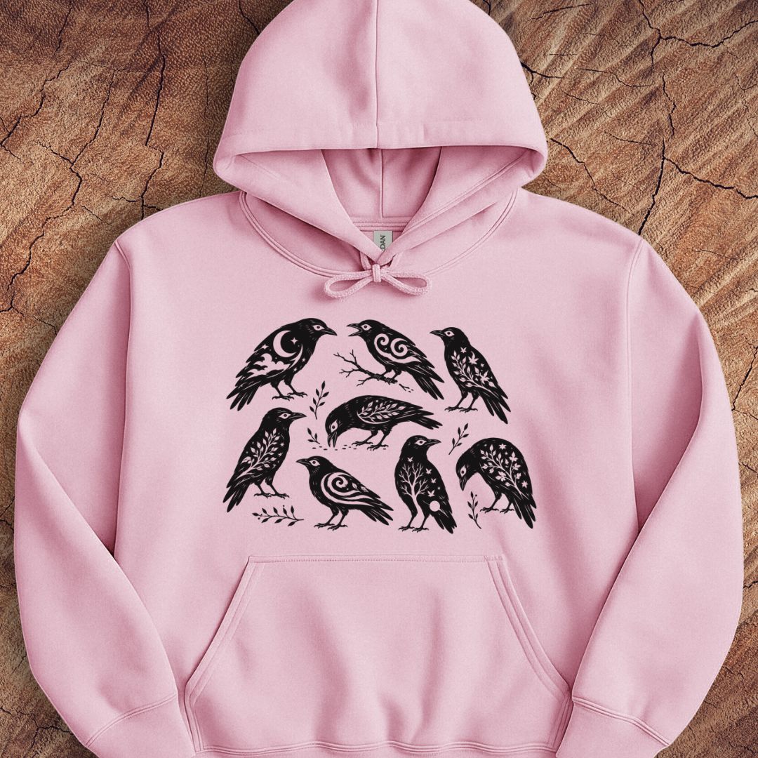 A light pink pullover hoodie with a black graphic print of seven stylized birds, laid flat on a textured wood background.