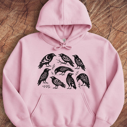 A light pink pullover hoodie with a black graphic print of seven stylized birds, laid flat on a textured wood background.