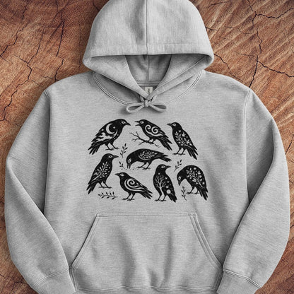 A light gray heather pullover hoodie featuring a black graphic print of seven stylized birds with intricate patterned feathers, laid flat on a textured brown wood background.