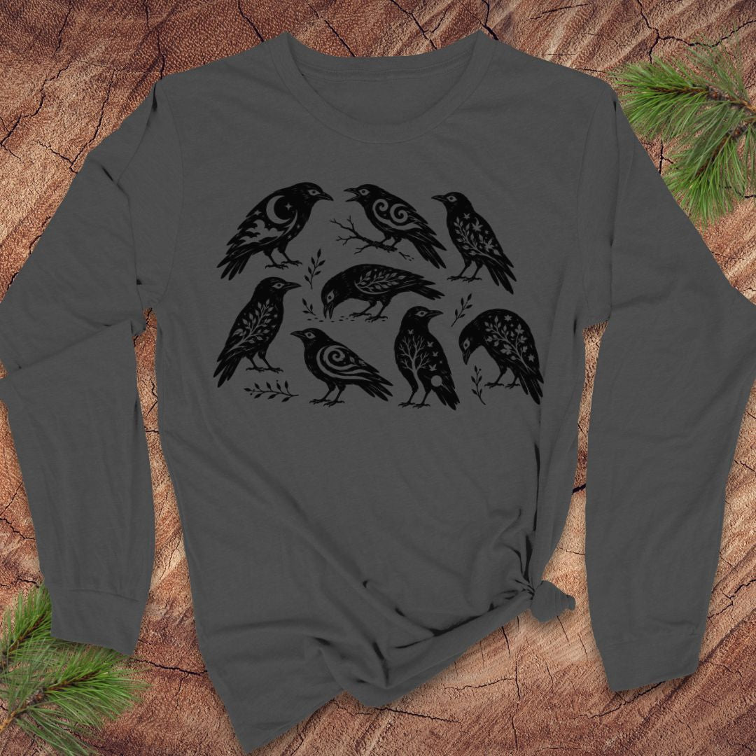 A dark gray long-sleeve t-shirt with a black graphic print of eight stylized crows, laid flat on a rustic wood background with pine branches in the corners.