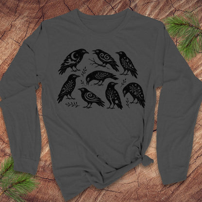 A dark gray long-sleeve t-shirt with a black graphic print of eight stylized crows, laid flat on a rustic wood background with pine branches in the corners.
