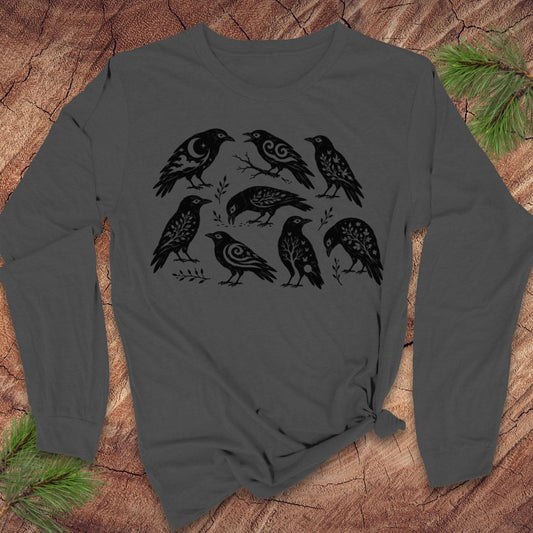 A dark gray long-sleeve t-shirt with a black graphic print of eight stylized crows, laid flat on a rustic wood background with pine branches in the corners.