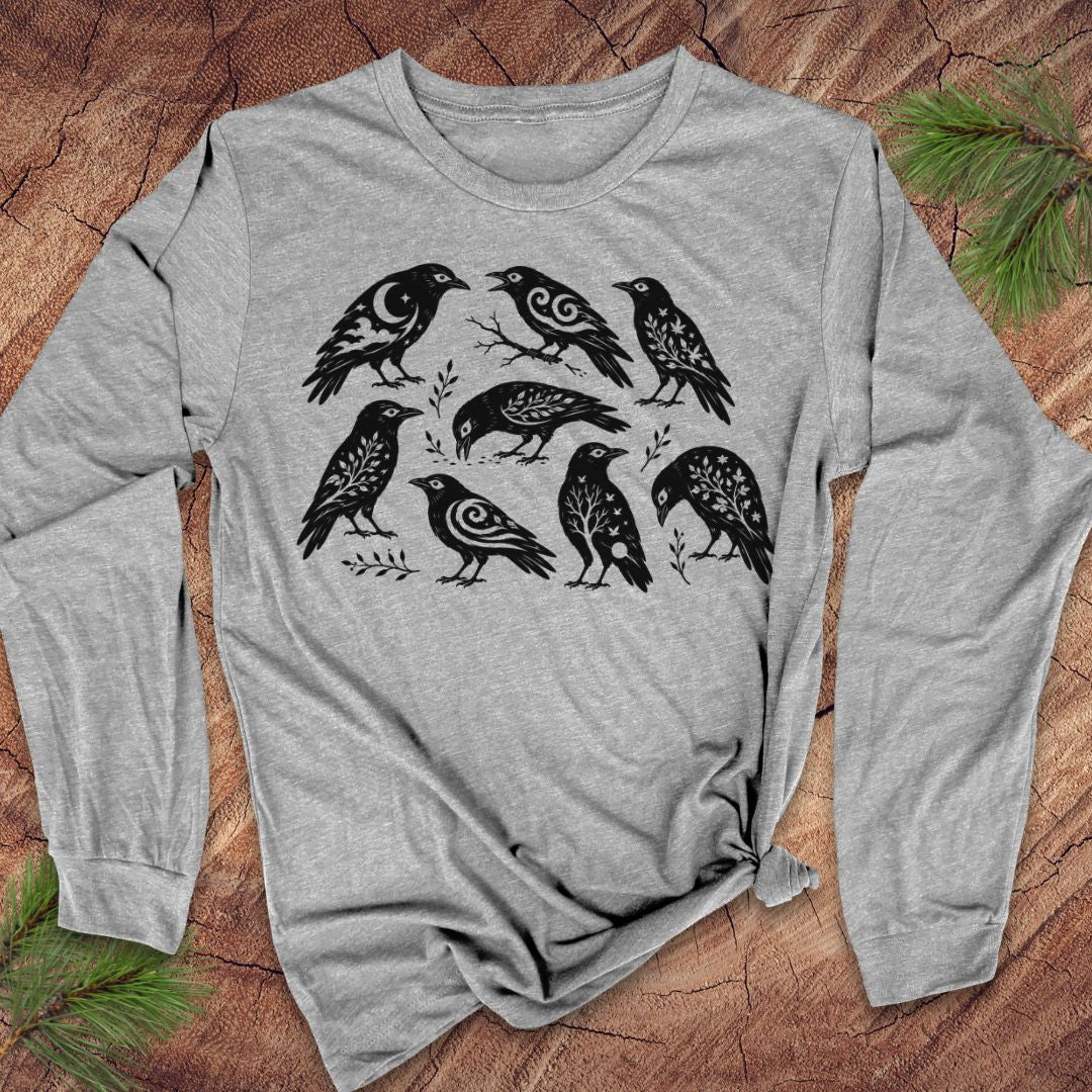 A heather gray long-sleeve t-shirt with a black graphic print of seven crows, laid flat on a rustic wood background with pine needles in the corners.