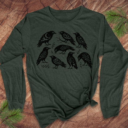 A dark forest green long-sleeve t-shirt with a black graphic print of eight stylized crows, laid flat on a rustic wood background with pine branches in the corners.