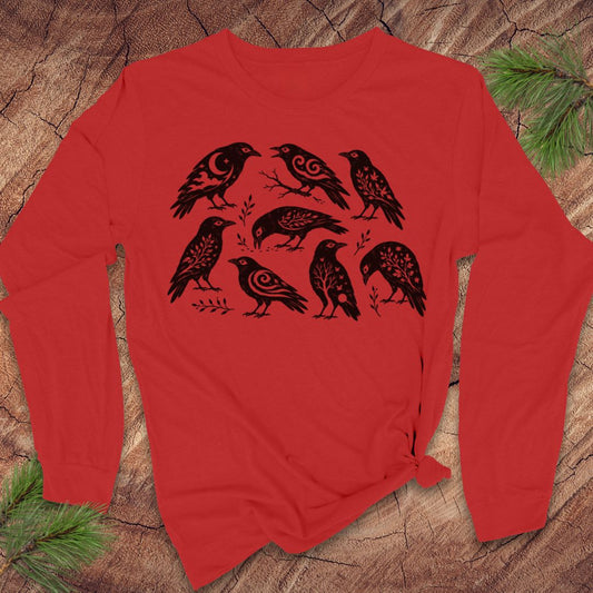 A red long-sleeve t-shirt with a black graphic print of seven stylized crows, laid flat on a rustic wood background with pine needles in the corners.