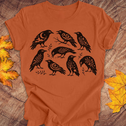 An orange crewneck t-shirt featuring a black graphic print of eight stylized crows with intricate patterned details, laid flat on a rustic wood background with autumn leaves.