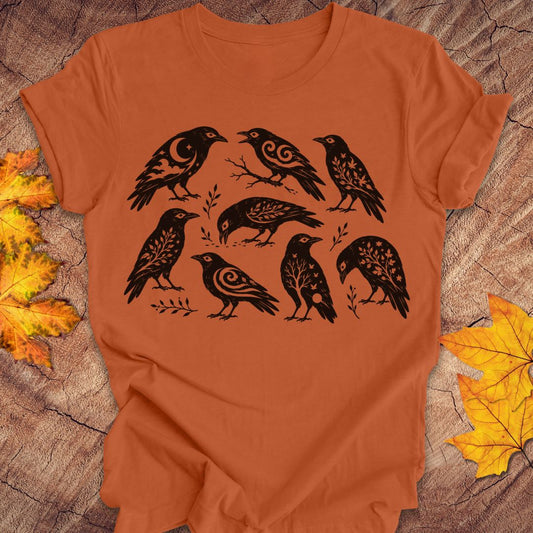 An orange crewneck t-shirt featuring a black graphic print of eight stylized crows with intricate patterned details, laid flat on a rustic wood background with autumn leaves.