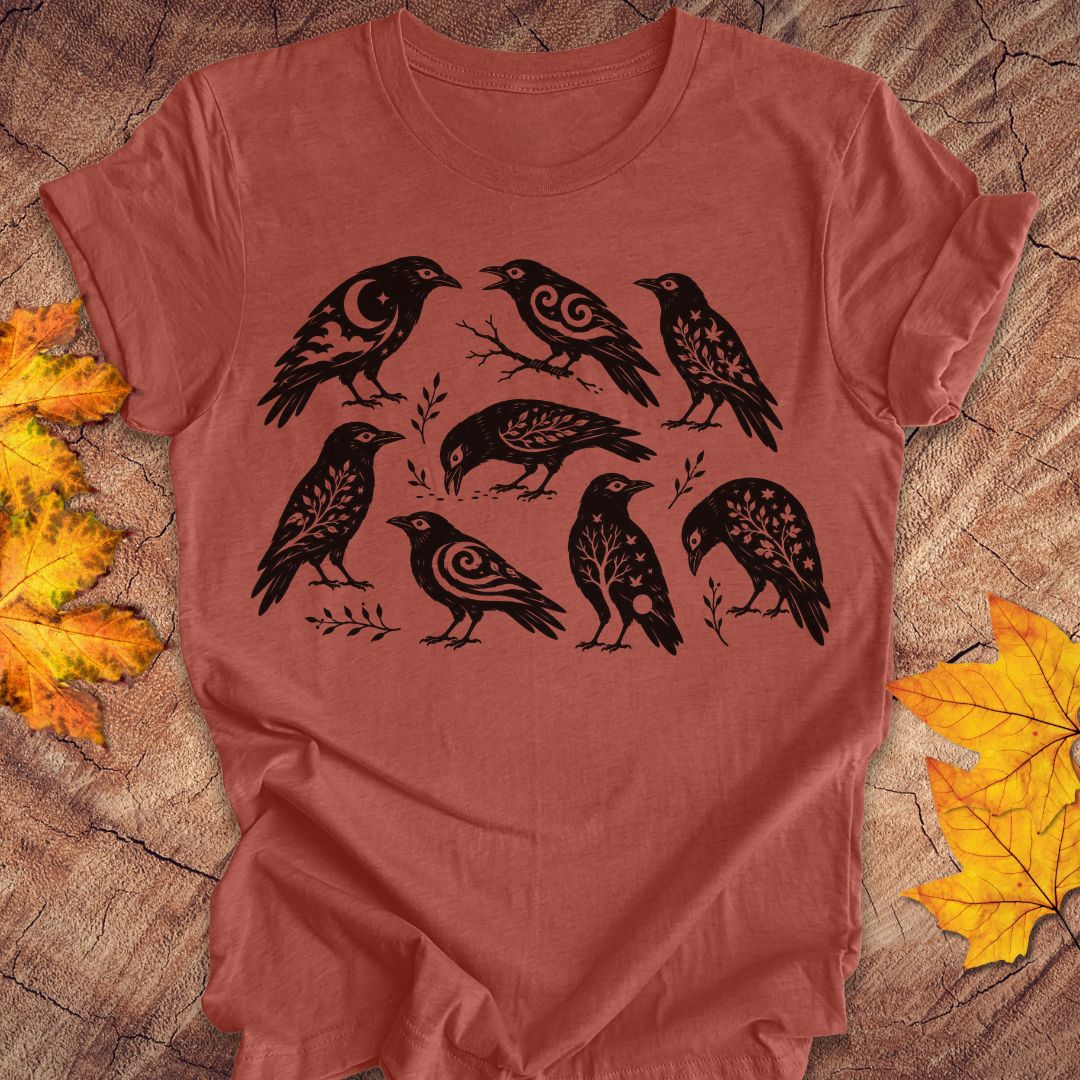 A terracotta-colored short-sleeve t-shirt with a black graphic print of eight stylized crows, laid flat on a rustic wood background with autumn leaves.
