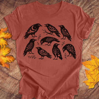 A terracotta-colored short-sleeve t-shirt with a black graphic print of eight stylized crows, laid flat on a rustic wood background with autumn leaves.