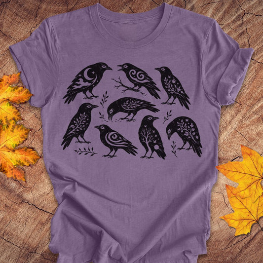 A lavender purple short-sleeve t-shirt with a black graphic print of eight stylized crows, laid flat on a rustic wood background with autumn leaves.