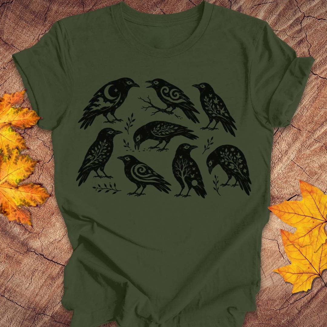 An olive green short-sleeve t-shirt with a black graphic print of eight stylized crows, laid flat on a rustic wood background with autumn maple leaves.