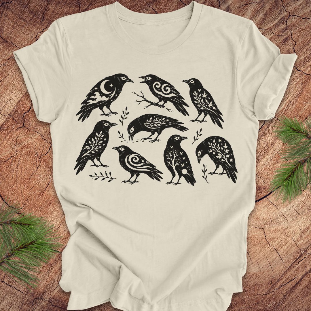 An off-white short-sleeve t-shirt with a black graphic print of eight stylized crows, laid flat on a rustic wood background with pine branches in the corners.