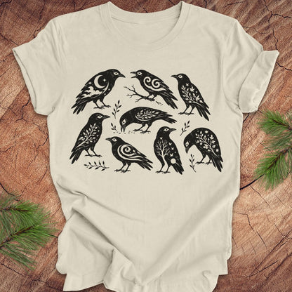 An off-white short-sleeve t-shirt with a black graphic print of eight stylized crows, laid flat on a rustic wood background with pine branches in the corners.