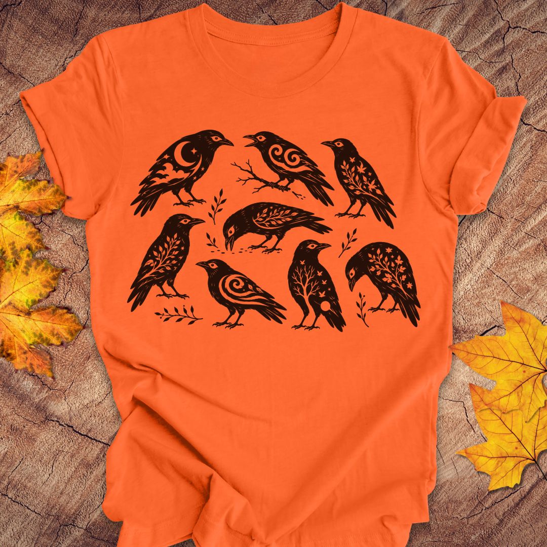An orange short-sleeve T-shirt laid flat on a rustic wood surface, decorated with crow illustrations and surrounded by yellow autumn leaves.