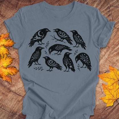A slate blue short-sleeve t-shirt with a black graphic print of eight stylized crows, laid flat on a rustic wood background with autumn leaves.