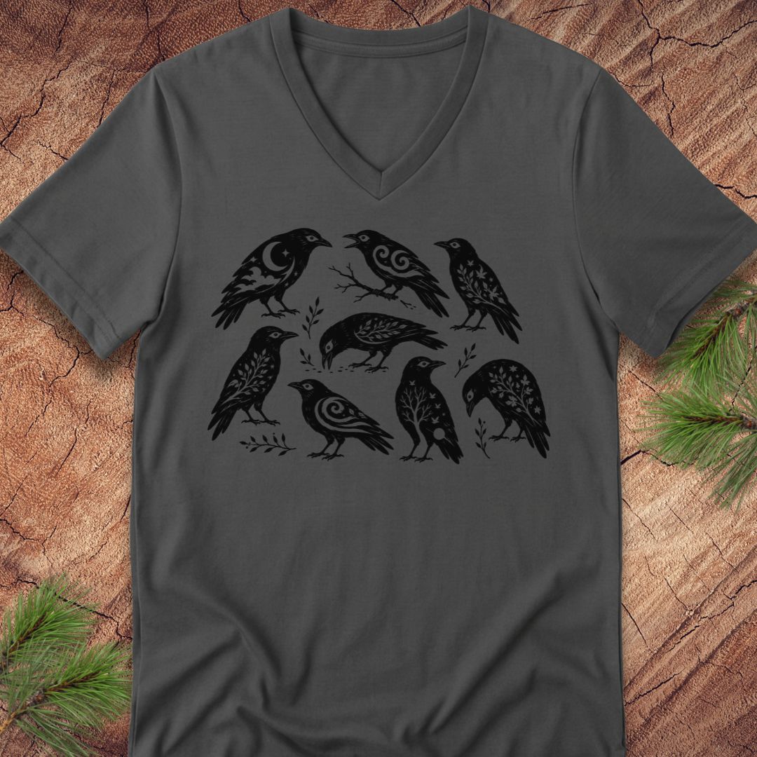 A dark gray v-neck t-shirt featuring a black graphic print of seven stylized birds, laid flat on a textured wood background with pine branches in the corners.