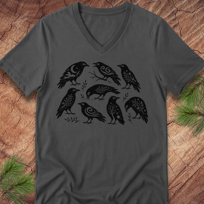 A dark gray v-neck t-shirt featuring a black graphic print of seven stylized birds, laid flat on a textured wood background with pine branches in the corners.