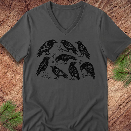 A dark gray v-neck t-shirt featuring a black graphic print of seven stylized birds, laid flat on a textured wood background with pine branches in the corners.