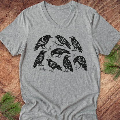 A heather gray V-neck t-shirt featuring a black graphic print of seven stylized birds, laid flat on a rustic wood background with pine branches in the corners.