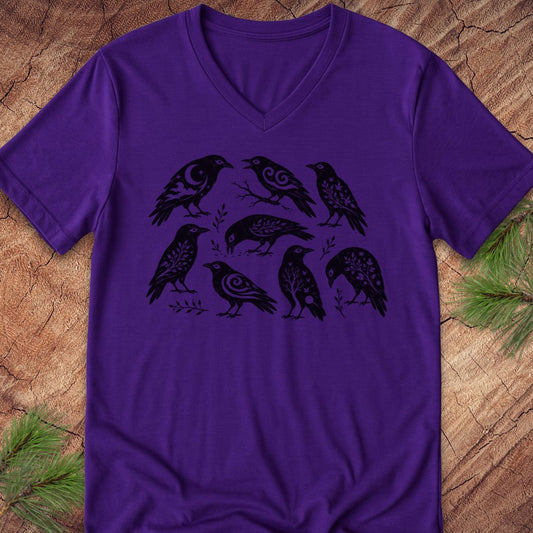 A purple short-sleeve V-neck t-shirt featuring a black graphic print of seven stylized birds with intricate patterned details, laid flat on a rustic wood background with pine needles in the corners.