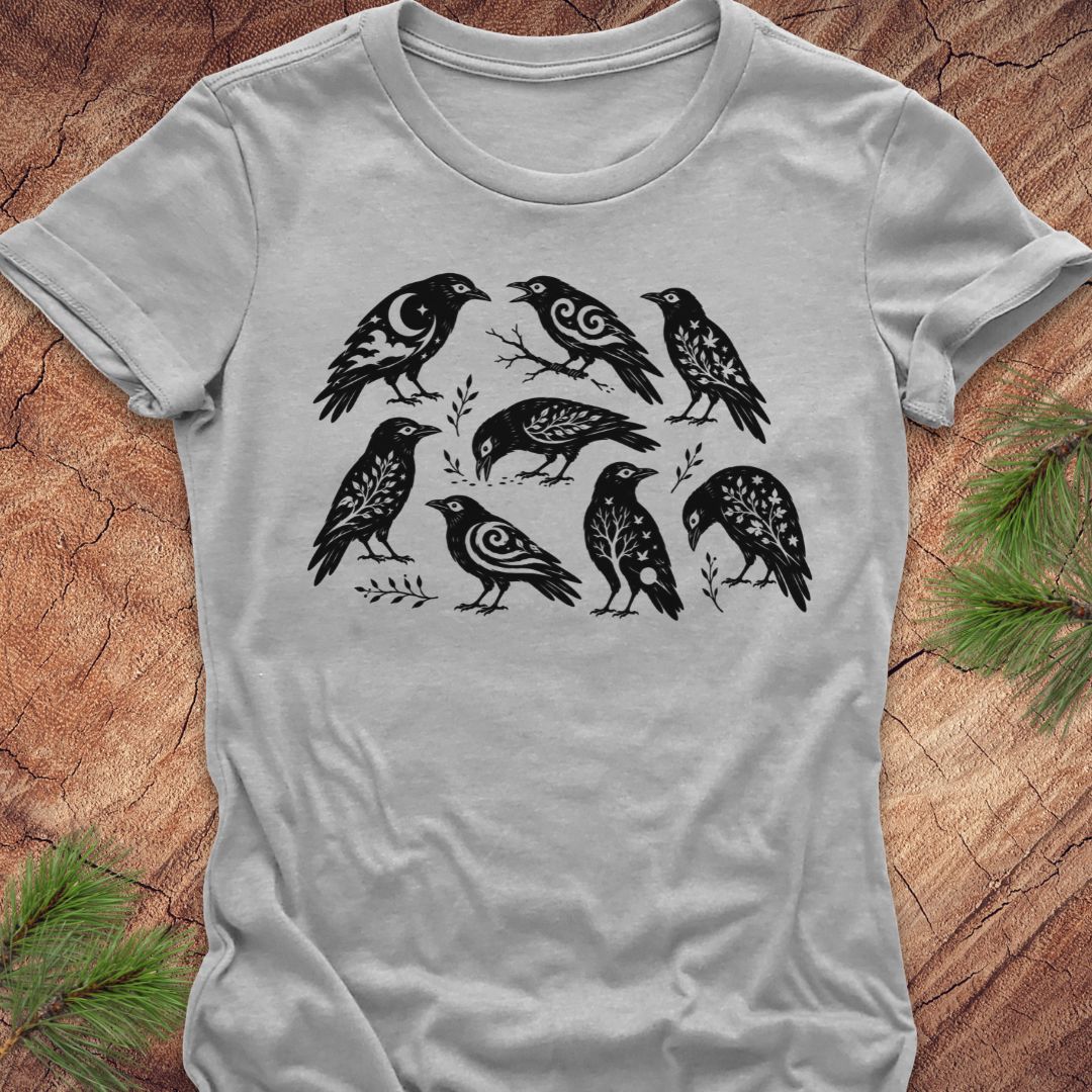 A light gray short-sleeve t-shirt with a black graphic print of seven stylized birds, laid flat on a rustic wood background with pine needles in the corners.