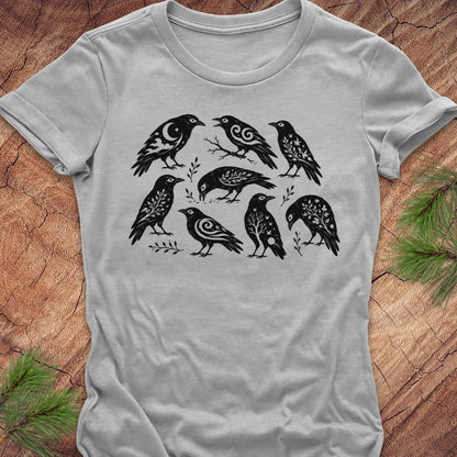 A light gray short-sleeve t-shirt with a black graphic print of seven stylized birds, laid flat on a rustic wood background with pine needles in the corners.