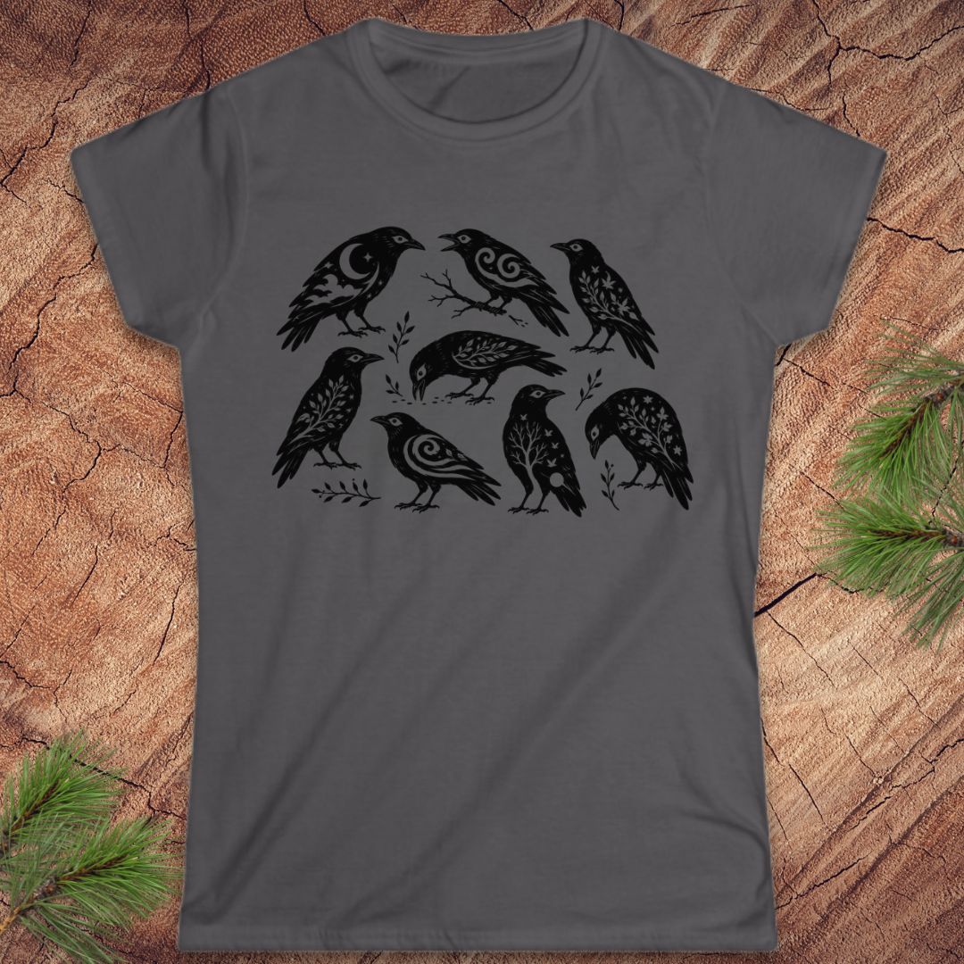 A dark gray short-sleeve t-shirt with a black graphic print of seven stylized birds, laid flat on a rustic wood background with pine branches in the corners.