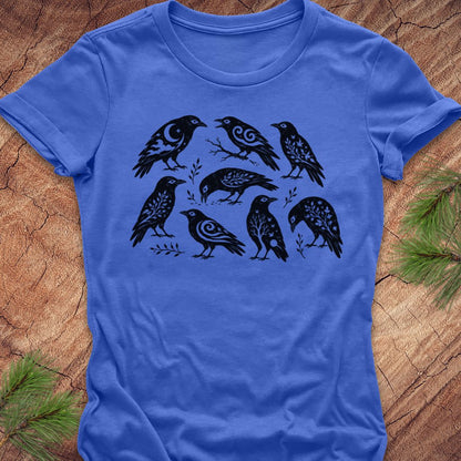 A royal blue short-sleeve t-shirt with a black graphic print of eight stylized birds arranged in two rows, laid flat on a textured wood background with pine needles in the corners.