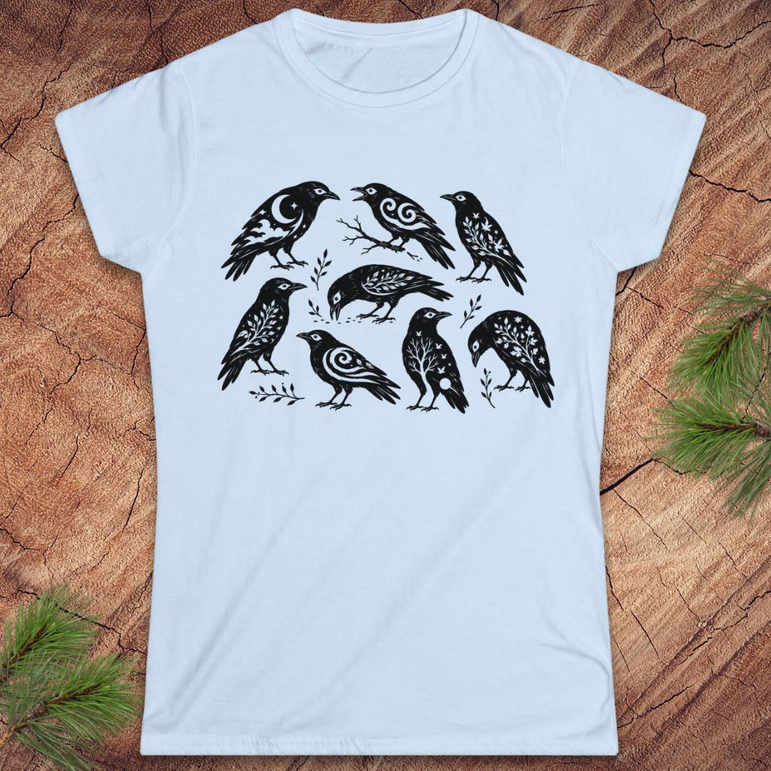 A light blue short-sleeve t-shirt with a black graphic print of eight stylized birds, laid flat on a rustic wood background with pine branches in the corners.