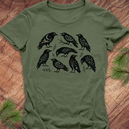 An olive green short-sleeve t-shirt with a black graphic print of seven stylized birds, laid flat on a rustic wood background with pine needles in the corners.