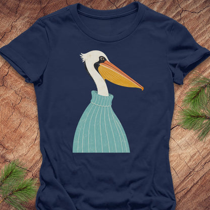 A navy blue short-sleeve t-shirt featuring a stylized graphic of a white pelican wearing a light blue striped sweater, laid flat on a textured wood background with pine branches in the corners.