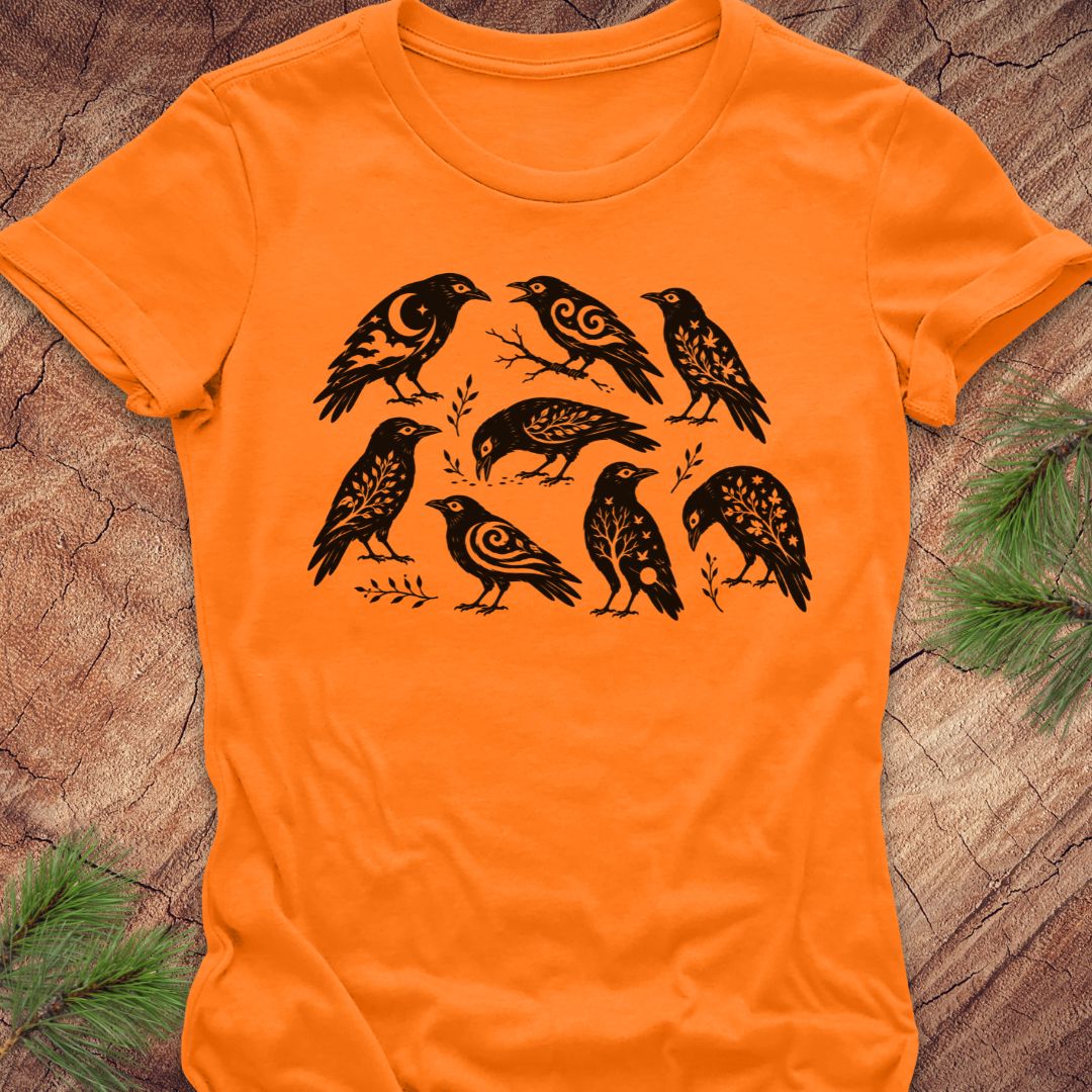An orange short-sleeve crewneck t-shirt featuring a black graphic print of seven stylized birds with intricate patterned details, laid flat on a rustic wood background with pine needles in the corners.