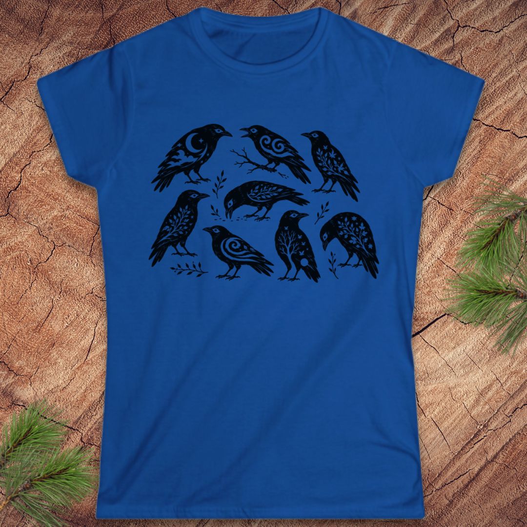 A royal blue short-sleeve t-shirt with a black graphic print of eight stylized ravens, laid flat on a textured wood background with pine branches in the corners.