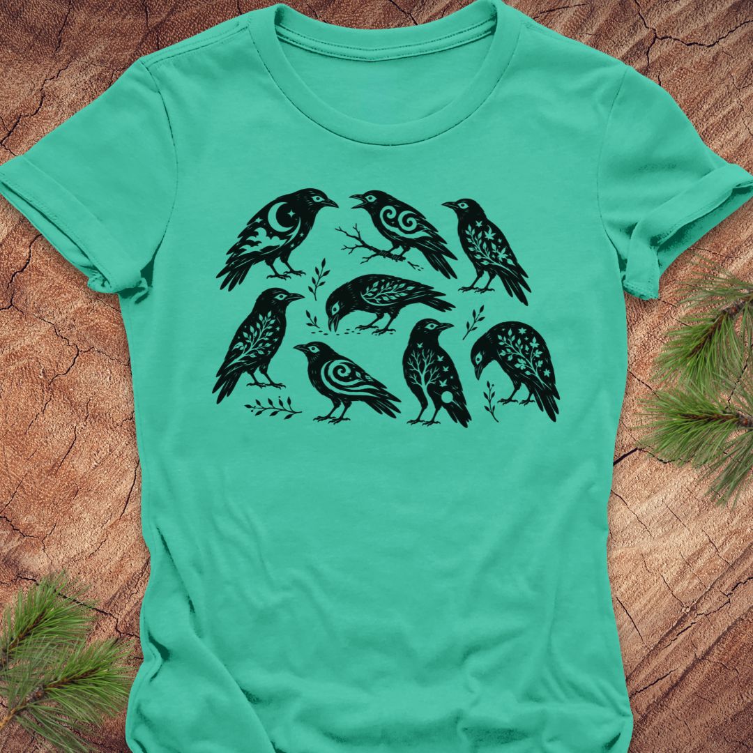 A mint green short-sleeve t-shirt with a black graphic print of seven stylized birds, laid flat on a textured brown wood background with pine needles in the corners.