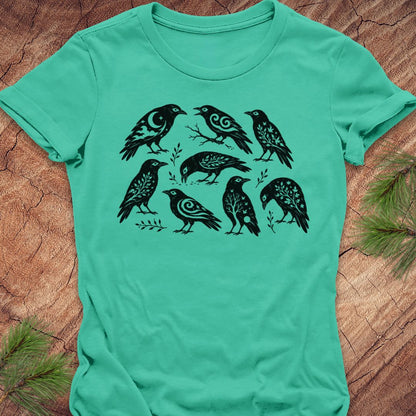 A mint green short-sleeve t-shirt with a black graphic print of seven stylized birds, laid flat on a textured brown wood background with pine needles in the corners.