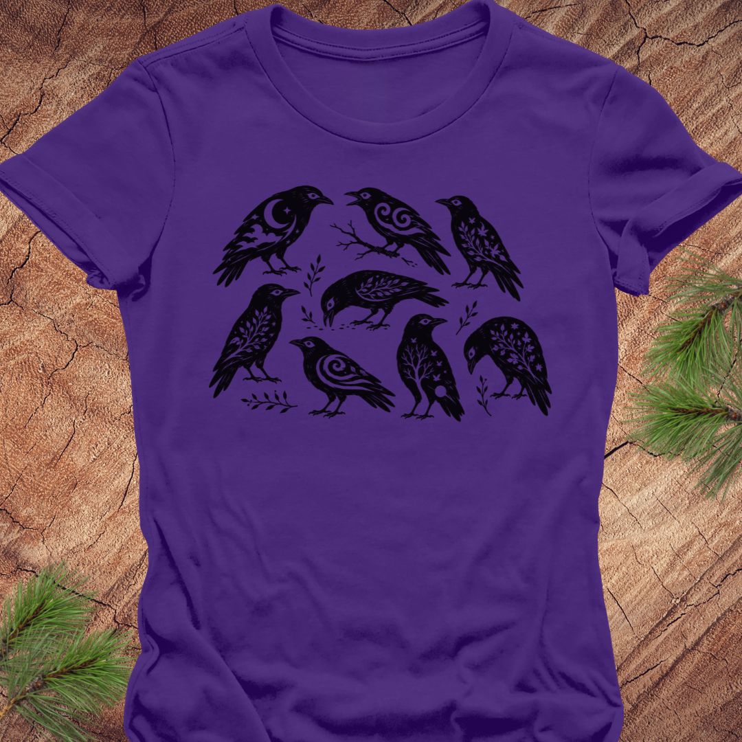 A purple short-sleeve crewneck t-shirt featuring a black graphic print of seven stylized birds on branches, laid flat on a rustic wood background with pine needles in the corners.