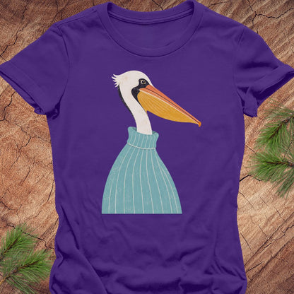 A purple short-sleeve crewneck t-shirt featuring a stylized graphic of a white pelican wearing a light blue striped sweater, laid flat on a rustic wood background with pine needles.