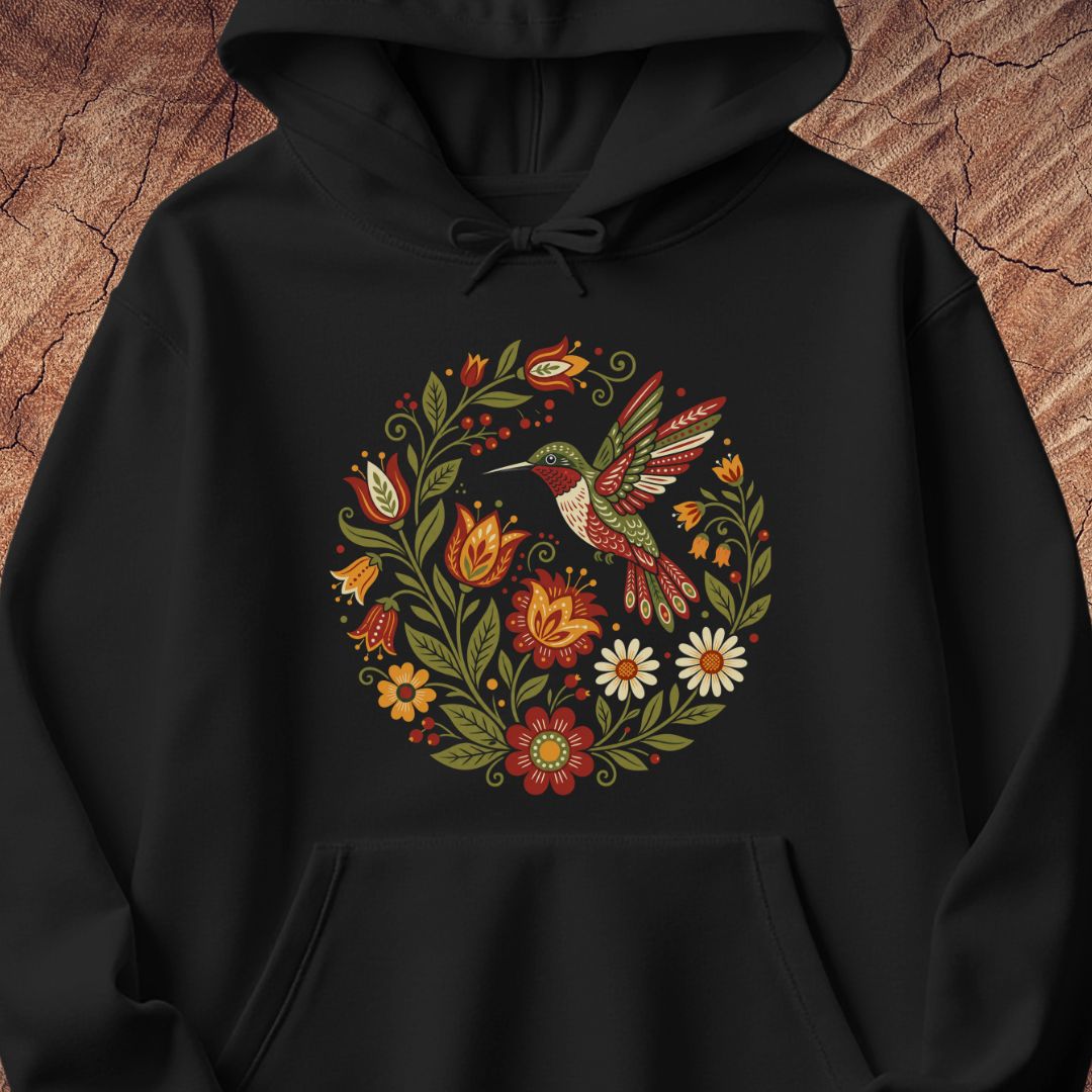 A black pullover hoodie featuring a colorful circular graphic of a hummingbird surrounded by flowers and leaves, displayed against a rustic wood background.