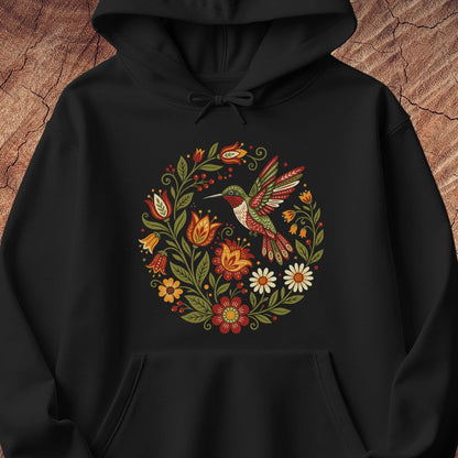 A black pullover hoodie featuring a colorful circular graphic of a hummingbird surrounded by flowers and leaves, displayed against a rustic wood background.