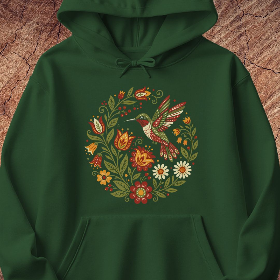 A dark green pullover hoodie featuring a colorful central graphic of a hummingbird surrounded by a circular wreath of flowers and leaves, displayed against a textured brown wood background.