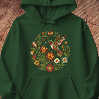 A dark green pullover hoodie featuring a colorful central graphic of a hummingbird surrounded by a circular wreath of flowers and leaves, displayed against a textured brown wood background.