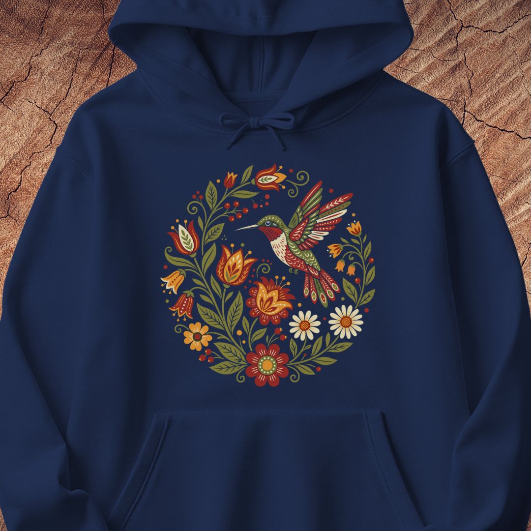 A navy blue pullover hoodie featuring a colorful folk-art style graphic of a hummingbird surrounded by flowers and leaves, displayed against a rustic wood background.