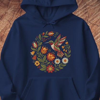 A navy blue pullover hoodie featuring a colorful folk-art style graphic of a hummingbird surrounded by flowers and leaves, displayed against a rustic wood background.