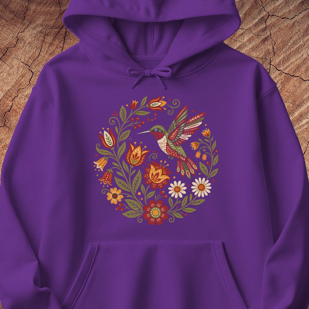 A purple pullover hoodie with a colorful circular graphic of a hummingbird surrounded by flowers, displayed flat against a rustic wood background.