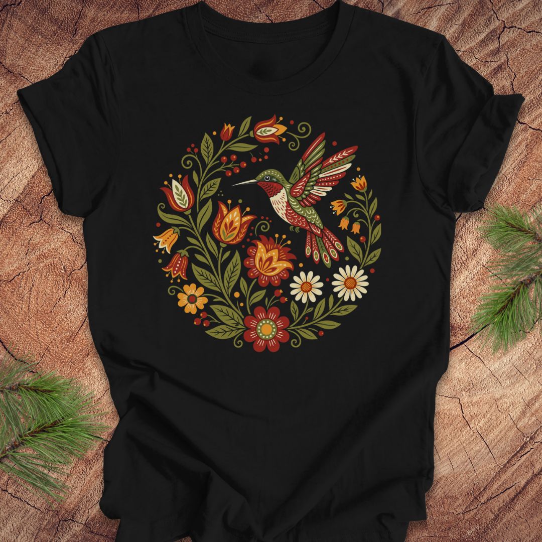 A black short-sleeve t-shirt with a colorful folk-art style graphic of a hummingbird and flowers, laid flat on a rustic wood background with pine branches in the corners.