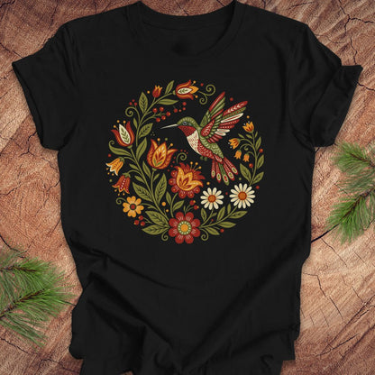 A black short-sleeve t-shirt with a colorful folk-art style graphic of a hummingbird and flowers, laid flat on a rustic wood background with pine branches in the corners.